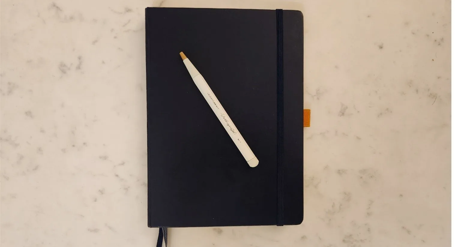 Closed dark blue hardcover notebook with an elastic band and an orange bookmark, a white Leuchtturm1917 Drehgriffel pen lying on the cover