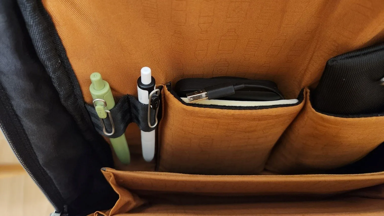 Front pocket of a bag with an orange lining: two pens clipped into elastic loops, a power bank and a USB cable in the compartment next to them