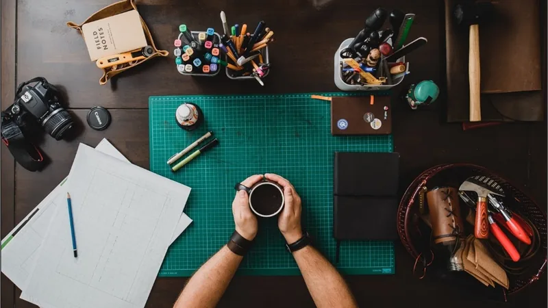 Top-down view of a desk: hands holding a mug on a green cutting mat, camera, markers, notebooks, leather tools and sheets of paper around