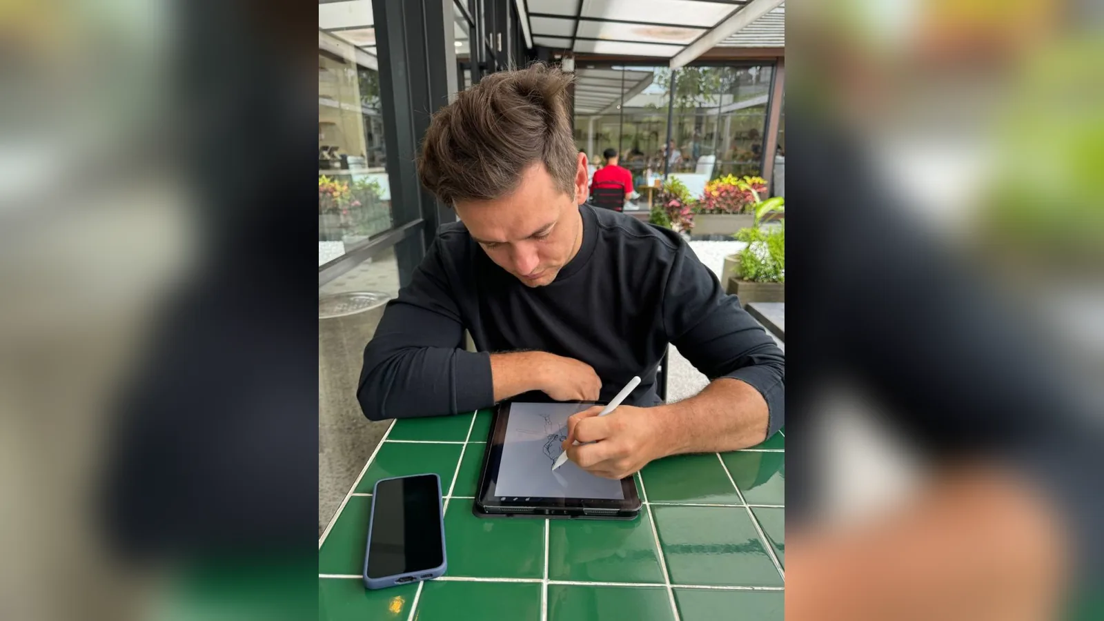 Maksim writing with a stylus on a tablet at a cafe table, phone beside him